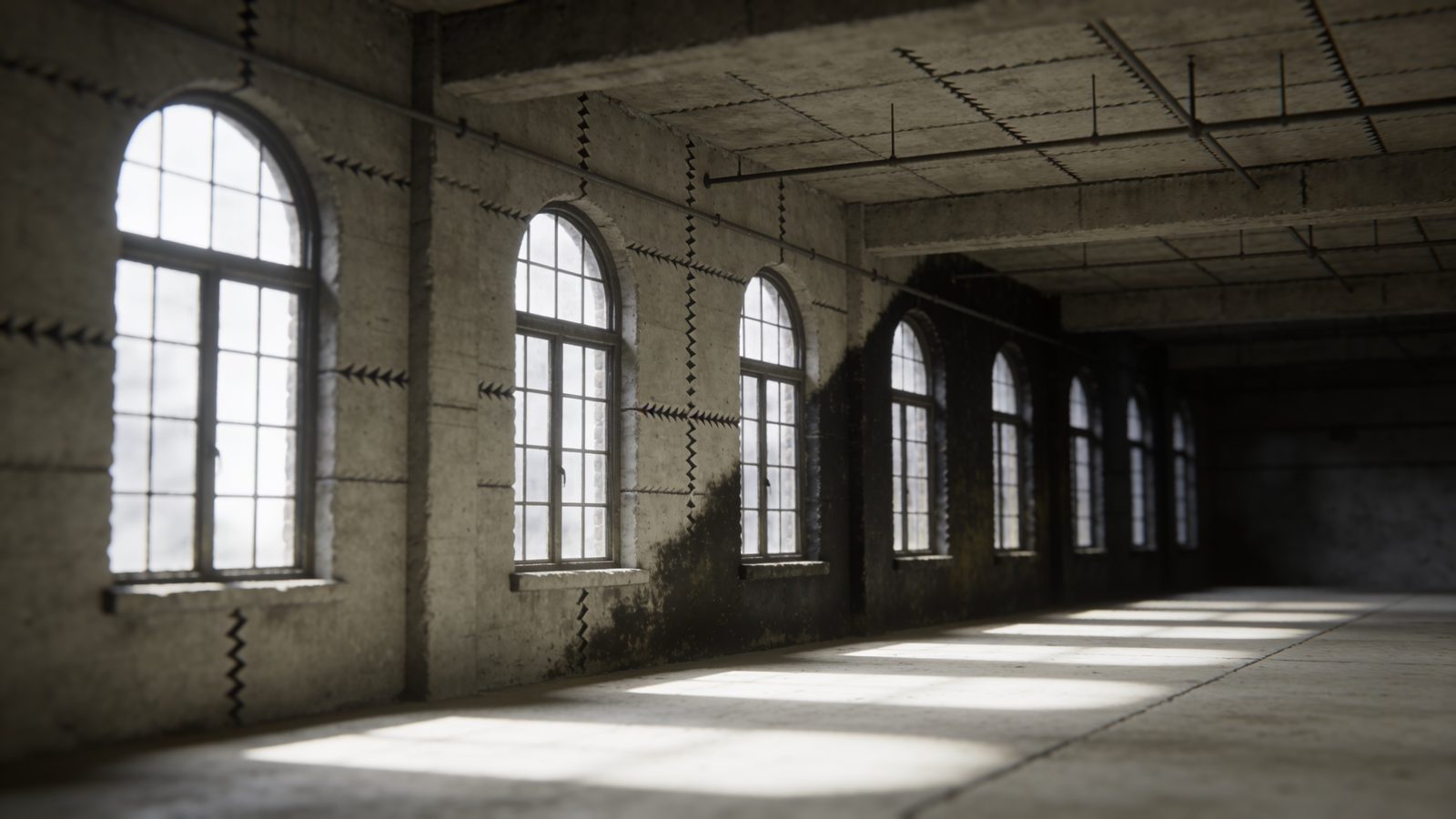 An old mill building floor at grade, coarse century-old concrete with a saw-cut joint and a damp shadow at the wall line, tall arched windows throwing light across it